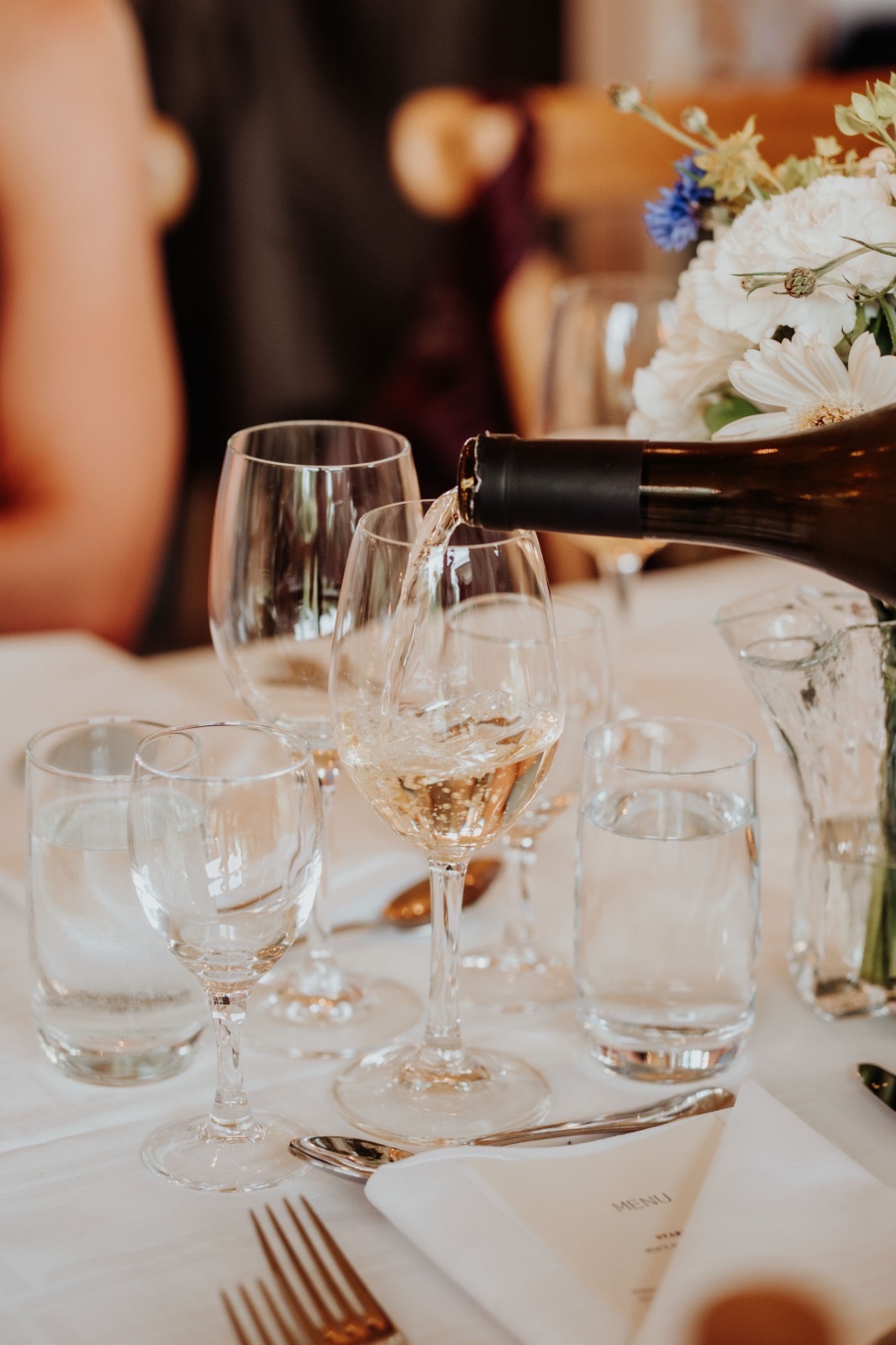 Vitt vin hälls upp i ett glas vid det dukade bordet med en blomsterbukett i bakgrunden.