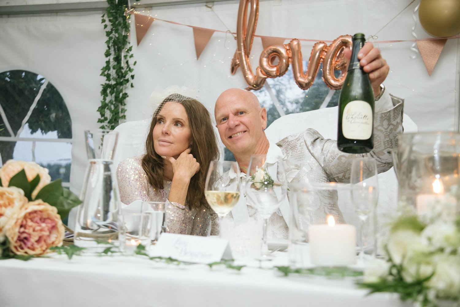 En man och en kvinna vid ett bröllopsbord, mannen håller upp en flaska champagne.