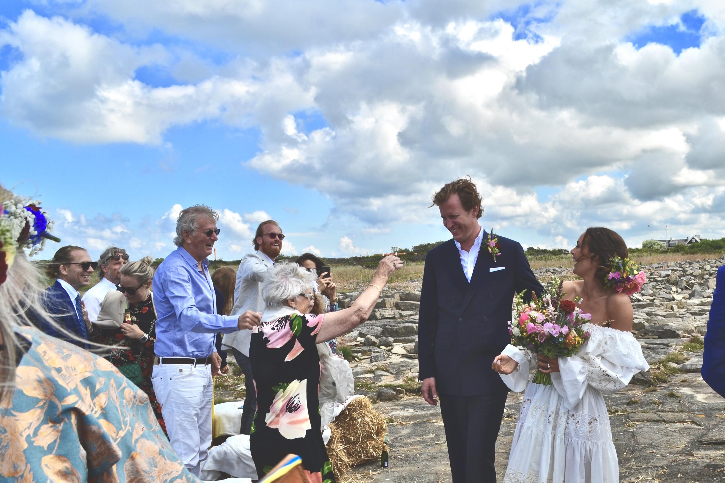 Brudpar på klipporna vid havet, omgivna av gäster som skålar.